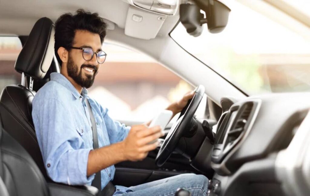 Happy young indian guy using smartphone while driving car, using navigation system gps. Eastern businessman chatting with friends or clients on mobile phone while going to office by car.